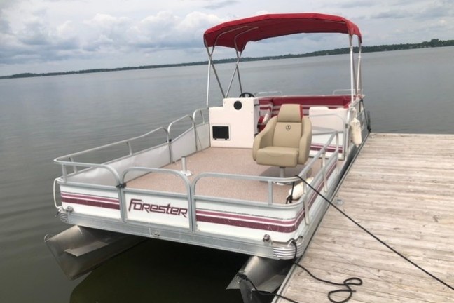 Pontoon boat docked at wooden pier with red canopy and calm water.