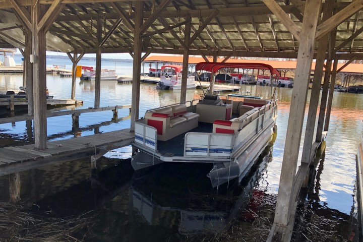 Pontoon boat docked under wooden boathouse on a lake.
